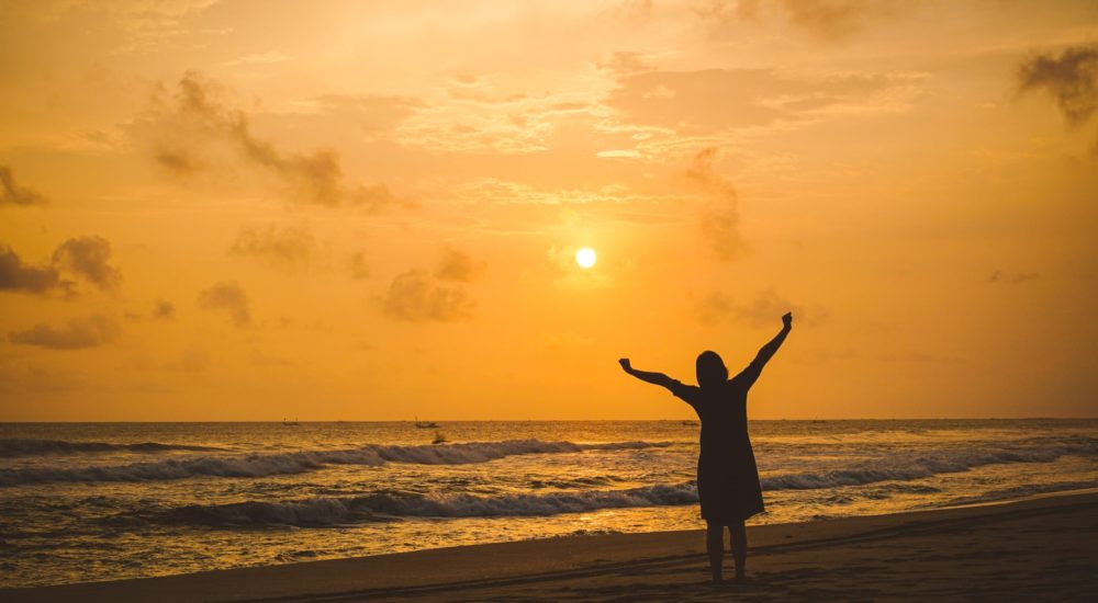 silhouette of woman with arms raised overhead against an orange sky during sunrise