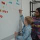 student writes on a white board while a teacher looks on with care